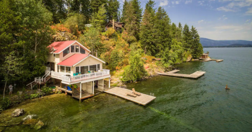 A scenic lakeside house with a red roof, surrounded by trees and docks on a calm water body under a blue sky.
