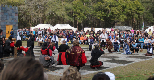 A crowd watches a medieval-themed performance on a large chessboard, with actors in period costumes engaging in a scene.
