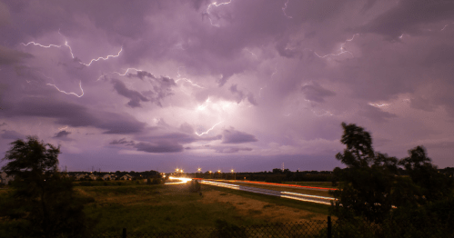 A dramatic sky filled with lightning over a highway, with streaks of light from passing vehicles and dark clouds.