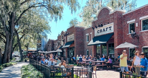 A lively outdoor dining area with people enjoying meals at a brick restaurant, surrounded by trees and sunny skies.