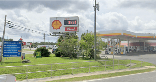 Gas station with a Shell sign displaying fuel prices, surrounded by greenery and a clear sky.