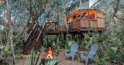 A cozy treehouse in the woods, featuring a fire pit and two chairs, surrounded by trees and string lights.