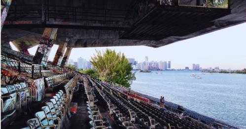 Abandoned stadium with graffiti, overlooking a waterfront and city skyline in the background.