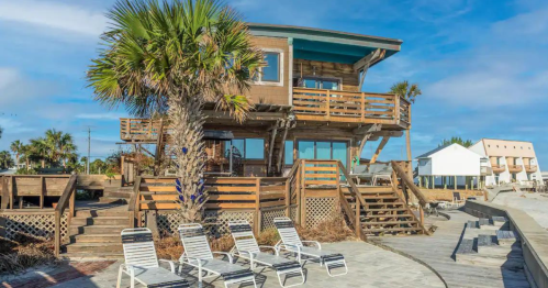 A wooden beach house with a palm tree, surrounded by lounge chairs and a boardwalk, under a blue sky.