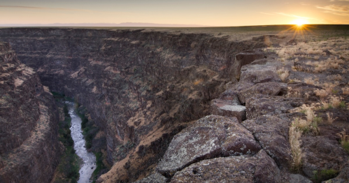A scenic view of a canyon at sunset, with a river winding through rocky cliffs and golden light illuminating the landscape.