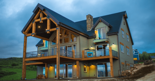 A modern mountain house with large windows, a wooden deck, and a stone chimney, set against a cloudy sky.