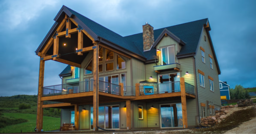 A modern mountain house with large windows, a wooden deck, and a stone chimney, set against a cloudy sky.