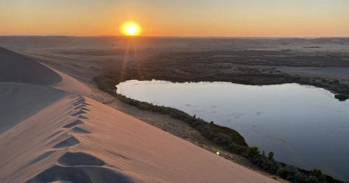 Sunset over a serene landscape, with a shimmering lake surrounded by rolling sand dunes and distant hills.