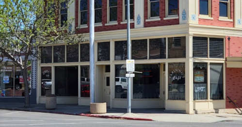 A corner café with large windows, featuring a brick exterior and trees nearby on a sunny day.