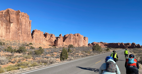 Cyclists riding on a road with stunning red rock formations and clear blue skies in the background.