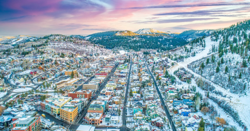 Aerial view of a snowy town surrounded by mountains, with colorful buildings and a winding road under a pastel sky.