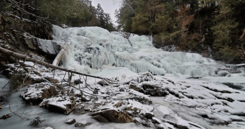 A frozen waterfall surrounded by snow-covered rocks and trees, creating a serene winter landscape.