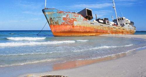 An abandoned, rusted ship stranded on a sandy beach with gentle waves lapping at its hull under a clear blue sky.