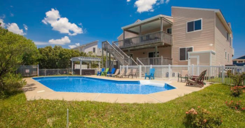 A sunny backyard with a pool, lounge chairs, and a two-story house surrounded by greenery and blue skies.