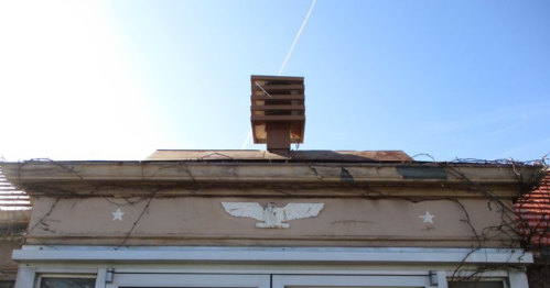 A weathered building facade with an eagle emblem, vines, and a wooden structure on the roof against a clear blue sky.