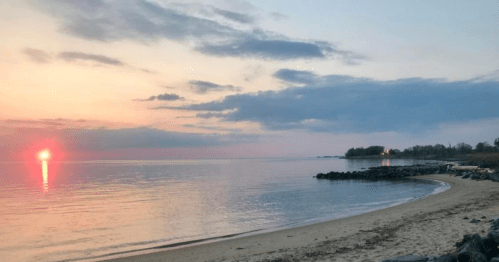 A serene beach at sunset, with calm waters reflecting the colorful sky and distant trees along the shore.