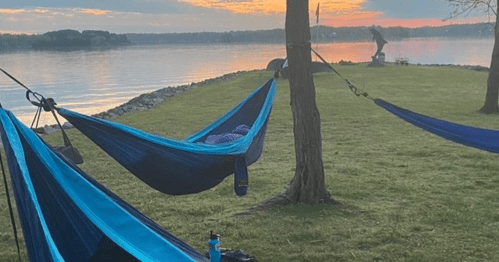 Two blue hammocks are set up by a calm lake at sunset, with a grassy area and trees in the background.