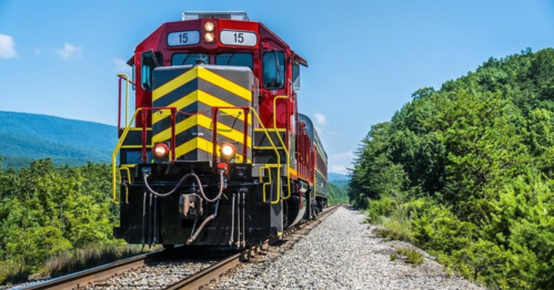 A bright red and yellow train on a track surrounded by green trees and mountains under a clear blue sky.