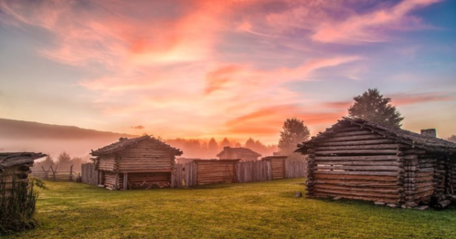 A serene landscape featuring rustic wooden cabins at sunrise, surrounded by mist and lush greenery.
