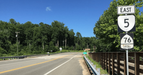 A road sign indicating East on Highway 5 and a bike route on Highway 76, surrounded by trees and clear blue skies.