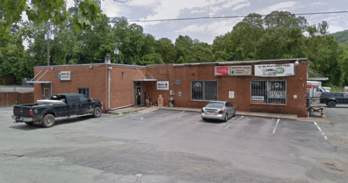 A brick building with signs, featuring a parking lot with a black truck and a silver car, surrounded by trees.