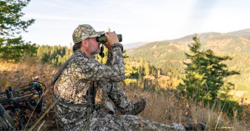 A hunter in camouflage gear uses binoculars to survey a mountainous landscape.
