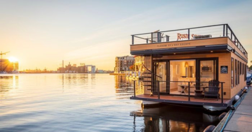 A modern houseboat at sunset, reflecting on calm waters with city skyline in the background.
