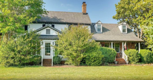 A charming two-story house surrounded by greenery and trees, featuring a front porch and a well-maintained lawn.