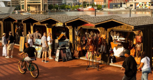 A bustling market with wooden stalls selling bags and crafts, people browsing, and a cyclist passing by.