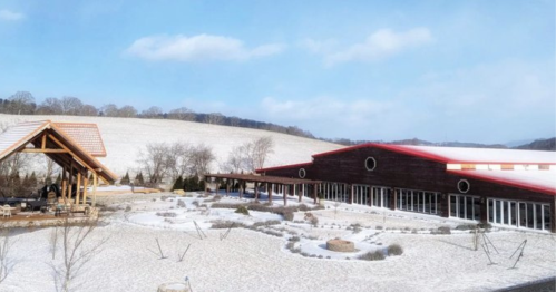 A snowy landscape featuring a modern building with a red roof and a wooden gazebo, surrounded by bare trees and hills.
