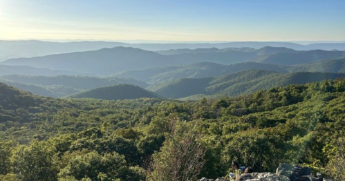 A panoramic view of rolling green mountains under a clear blue sky, with layers of hills fading into the distance.