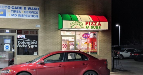 A red car parked outside a pizza and subs shop with a colorful awning, illuminated at night.