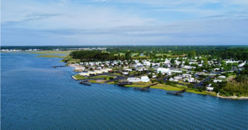 Aerial view of a coastal area with homes, docks, and greenery along the water's edge under a clear blue sky.