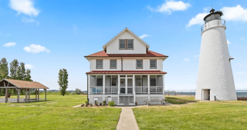 A white lighthouse beside a historic house, set against a clear blue sky and green grass.