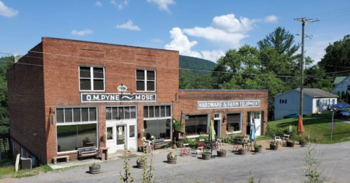 A brick storefront with "Q.M. Pyne" and "Hardware & Farm Equipment" signs, surrounded by greenery and blue skies.