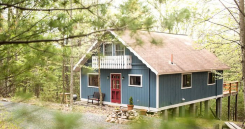 A charming blue cabin nestled in a wooded area, featuring a red door and a small porch.