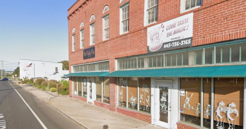 A brick building with large windows and green awnings, featuring signs for local businesses along a street.