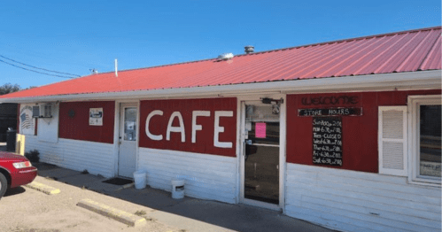 A small café with a red roof and white exterior, featuring a large "CAFE" sign and store hours posted on the door.