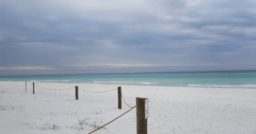 A serene beach scene with white sand, turquoise water, and a cloudy sky, bordered by wooden posts and rope.