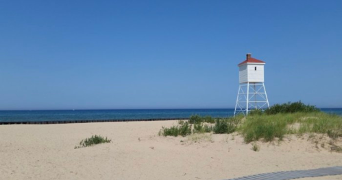 A white lighthouse on a sandy beach with a clear blue sky and calm ocean in the background.