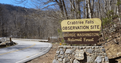 Sign for Crabtree Falls Observation Site in George Washington National Forest, with a winding road in the background.