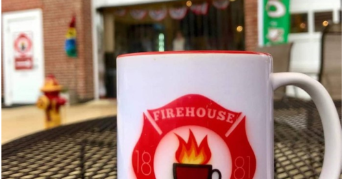 A close-up of a coffee mug with a firehouse logo, placed on a table outside a building decorated for a celebration.