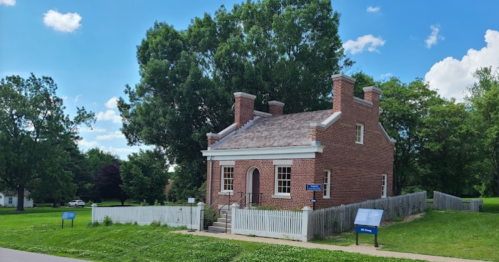 A historic brick house surrounded by greenery, with a white picket fence and informational signs nearby.