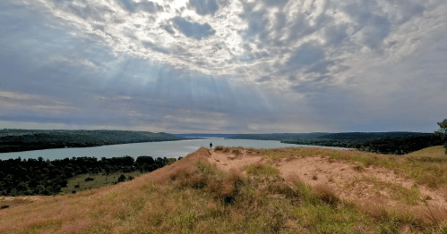 A scenic view of a river surrounded by hills under a cloudy sky, with sunlight breaking through the clouds.