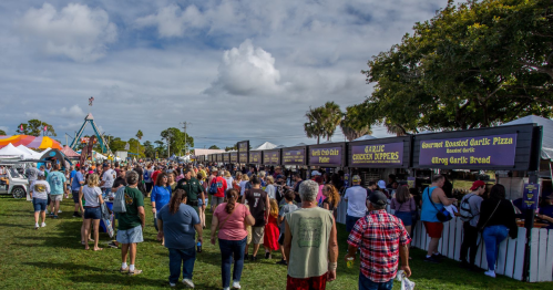A bustling outdoor festival with food stalls and a large crowd enjoying the event under a partly cloudy sky.