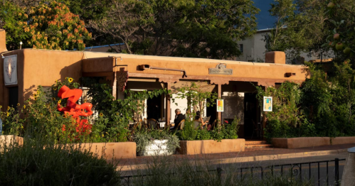 A charming adobe-style restaurant with greenery and colorful flowers, set against a sunny backdrop.