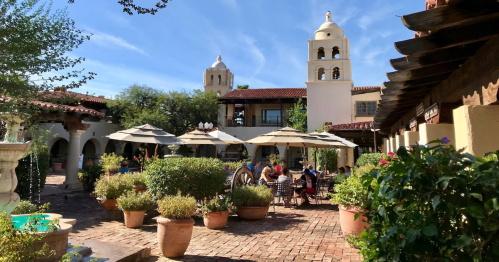 A sunny courtyard with potted plants, umbrellas, and people dining at tables, surrounded by Spanish-style architecture.