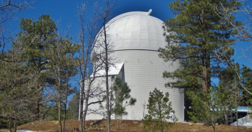 A large white observatory dome surrounded by pine trees under a clear blue sky.