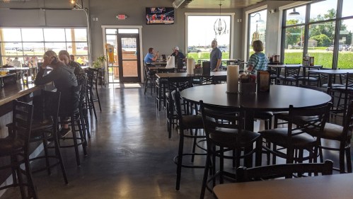 Interior of a casual dining restaurant with tables, bar seating, and patrons enjoying their meals. Large windows visible.