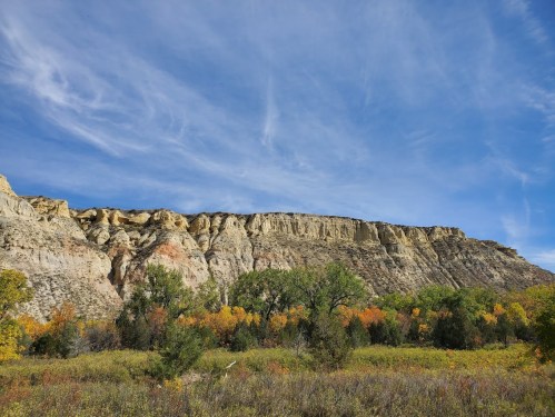 A scenic view of colorful autumn foliage in front of a rocky cliff under a blue sky with wispy clouds.